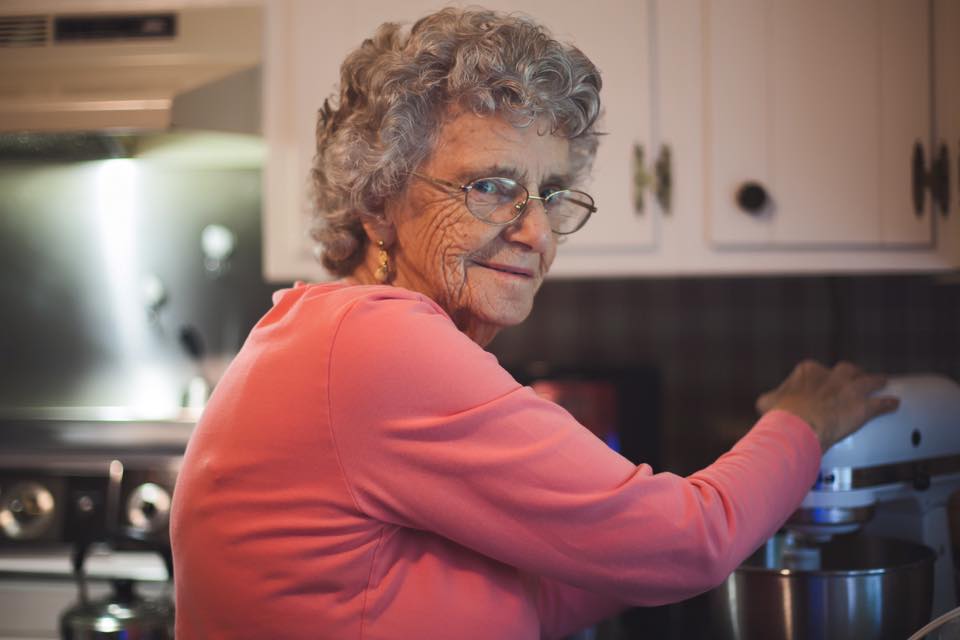 Elderly woman Grandma Sergie in a pink sweater smiles warmly while using a mixer in a cozy kitchen, evoking a sense of nostalgia and home cooking.
